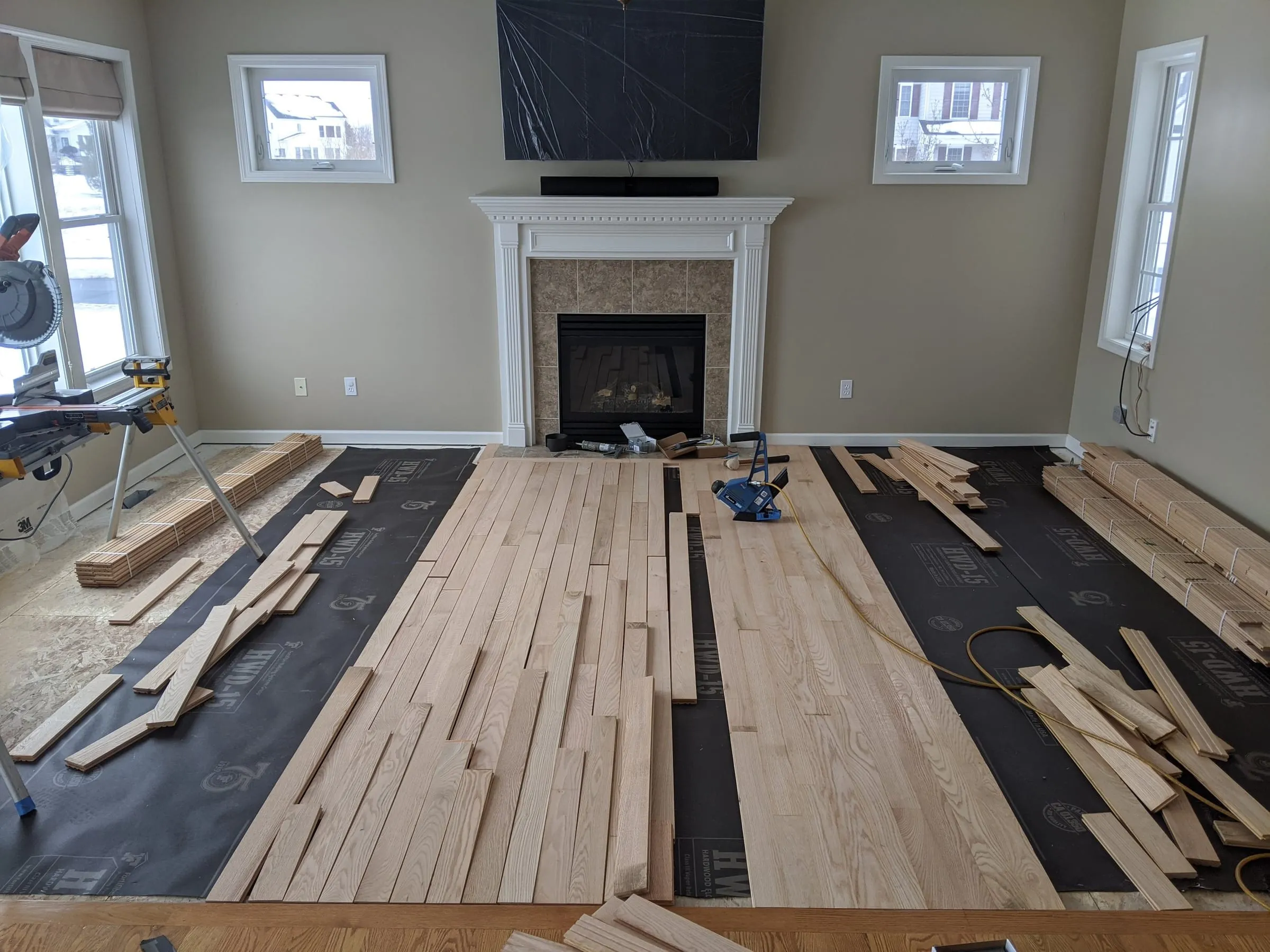 New oak boards being laid over underlayment in a living room with a fireplace